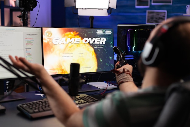 Speler die videogames streamt met een headset en verliest op de computer. Man kijkt naar monitoren met livestreamchat en online game, speelt en zendt gameplay uit met microfoon.