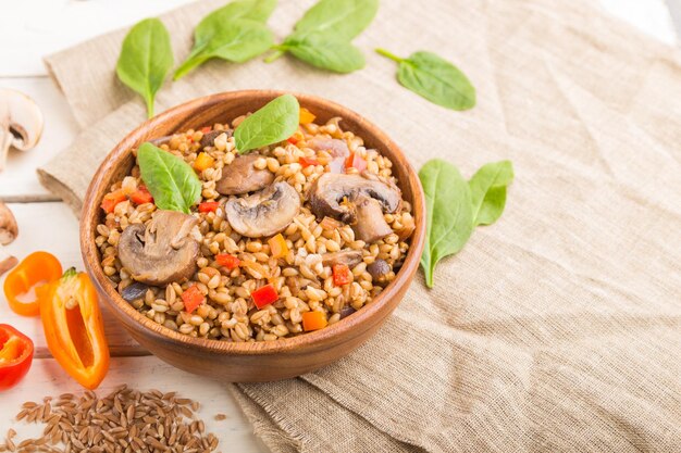 Spelt (dinkeltarwe) pap met groenten en champignons in houten kom op een witte houten ondergrond en linnen textiel. Zijaanzicht, selectieve aandacht. Russische traditionele keuken.