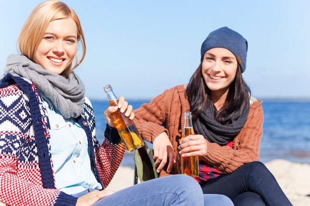 Trascorrere del tempo insieme. due belle giovani donne che tengono bottiglie di birra e sorridono seduti insieme sulla spiaggia