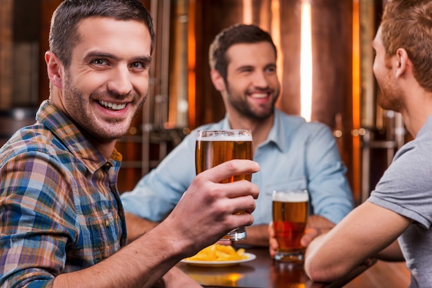 Проводить время с друзьями. Красивый молодой человек жарит пивом и улыбается, сидя со своими друзьями в пивном пабе