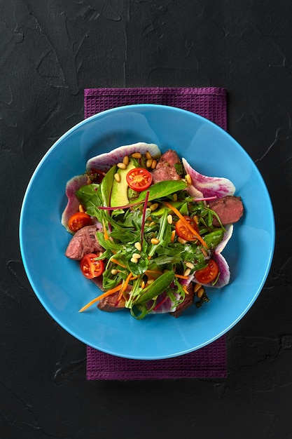 Spicy beef slices meat salad with tomatoes, parsley, radish and salad leaves spinach on black texture background table. Still life