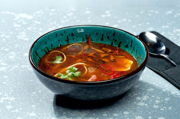 Spicy pan-asian ramen soup with noodles, meat, hot peppers and cilantro in a blue bowl on a gray surface