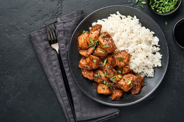 spicy teriyaki chicken fillet pieces with rice, green onions and black sesame seeds on black plate.