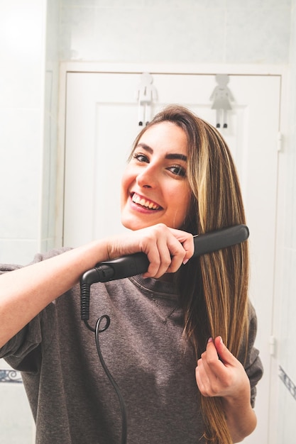 Spiegelstandpunt van een blanke jonge vrouw die haar haar rechttrekt met een stijltang in de badkamer.