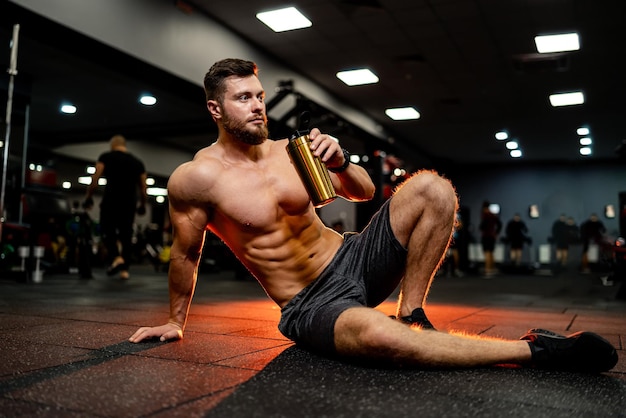 Spiervormige man op sportschool ontspannen drinken van eiwitcocktail Guy zittend op de vloer in moderne sportschool