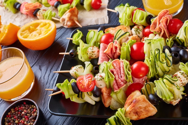 Spiesjes met kippenvlees, courgette, tomaten, mozzarella balletjes, plakjes salami, olijven op een zwarte plaat op een houten tafel met sinaasappelsap in glazen bekers, zomerpicknick recept, close-up