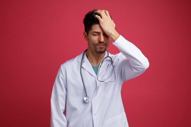 Spijtig jonge mannelijke arts dragen medisch uniform en stethoscoop om zijn nek, hand op het hoofd houden met gesloten ogen geïsoleerd op rode achtergrond