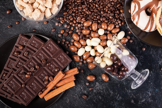 他のキャンディーの隣のスタジオ写真の暗い木製の背景にチョコレートのコーヒー豆とピーナッツとこぼれたグラス