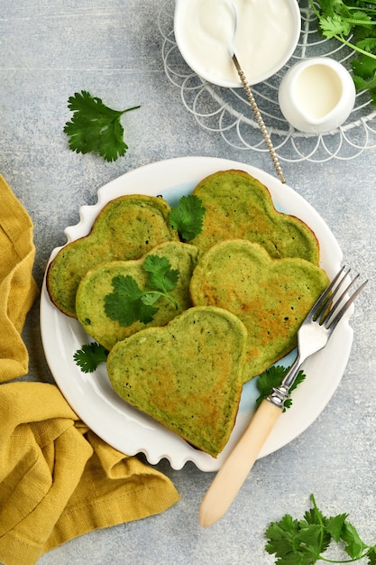 Spinach avocado pancakes in the shape of a heart with greek yogurt sauce and cherry tomatoes on white plate over light grey background. Breakfast food concept. Top view with copy space.