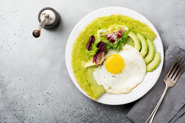 Spinach green crepes (pancakes) with fried egg, avocado and leaves of mix of salad on ceramic plate on gray concrete table. conceptof healthy breakfast. Selective focus. Top view. Copt space.