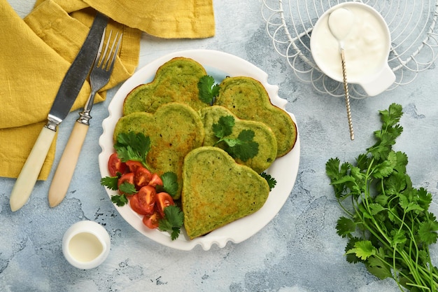 Spinach green Pancakes with greek yogurt sauce and cherry tomatoes in shape of heart over light grey background. Valentines day breakfast table. Love concept. Top view.