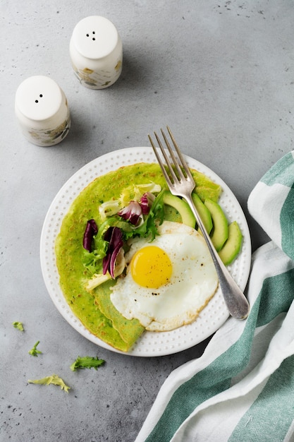 Spinazie groene pannenkoeken (pannenkoeken) met gebakken ei, avocado en bladeren van mix van salade op keramische plaat op grijs betonnen oppervlak. bovenaanzicht. koperen ruimte.