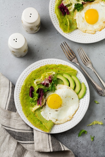 Spinazie groene pannenkoeken (pannenkoeken) met gebakken ei, avocado en bladeren van mix van salade op keramische plaat op grijs betonnen oppervlak. Bovenaanzicht. Koperen ruimte.
