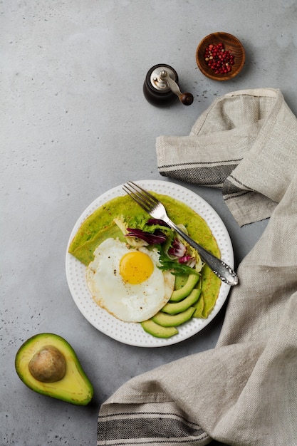 Spinazie groene pannenkoeken (pannenkoeken) met gebakken ei, avocado en bladeren van mix van salade op keramische plaat op grijs betonnen oppervlak. bovenaanzicht. koperen ruimte.