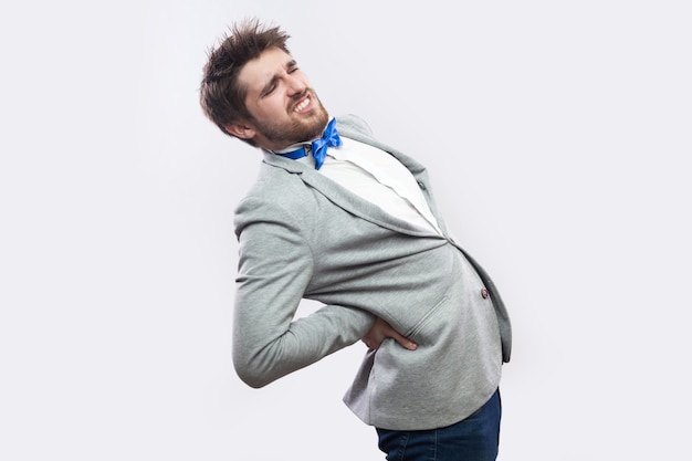 Spine or kidney pain. Profile side view portrait of young bearded businessman in casual grey suit and blue bow tie standing and holding his painful back. studio shot, isolated on light grey background