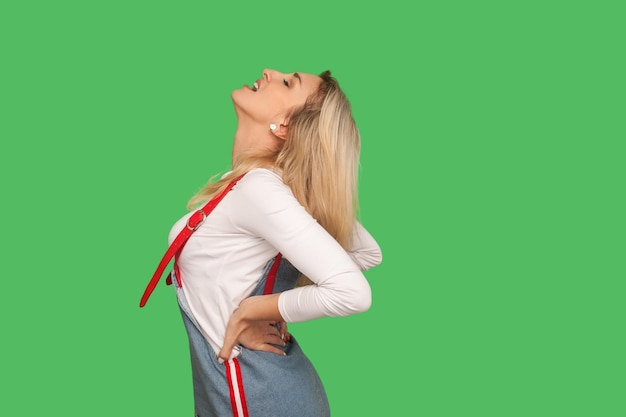 脊椎の問題デニムのオーバーオールに身を包んだ不健康な成人女性の肖像画が、緑の背景に分離された神経のピンチ神経をつまんだ急性腰痛に苦しんでいます。