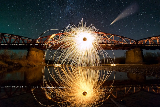 Spinning steel wool in abstract circle, firework showers of bright yellow sparks on long bridge reflected in river water under dark night starry sky.