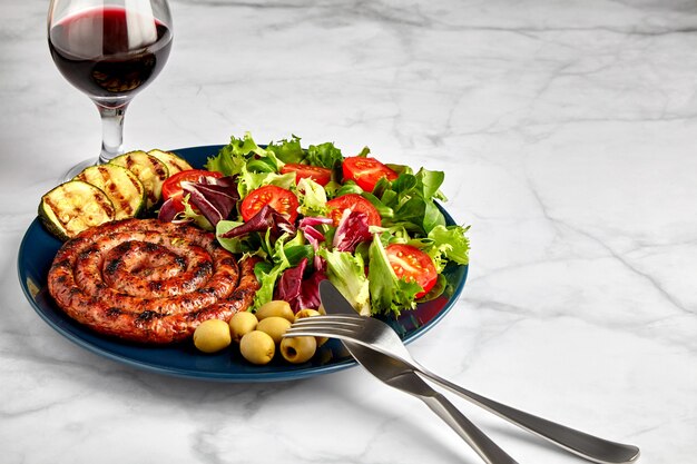 カトラリーと大理石のテーブルの上の赤ワインのグラスとプレート上の野菜とハーブとスパイラルグリルソーセージ