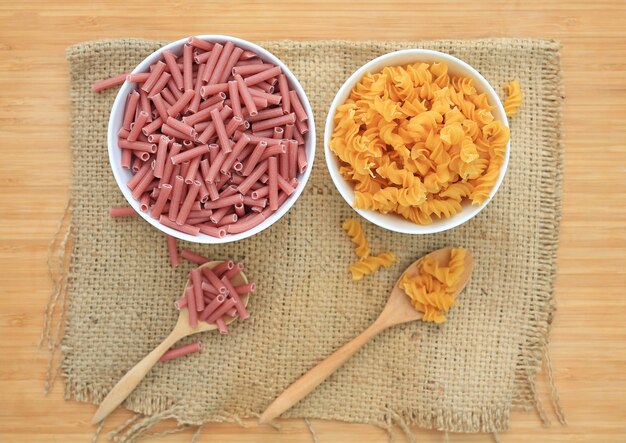 Spiral Macaroni and red pasta in white bowl on sack against wood.