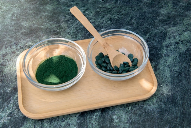 Foto spirulina poeder en pillen in glazen kommen op een houten dienblad met een houten lepel op een groene achtergrond