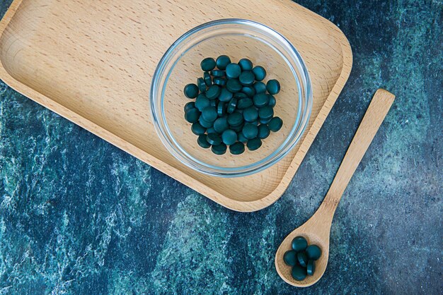 Foto compresse di spirulina in una ciotola di vetro e un cucchiaio di legno su un vassoio di legno su uno sfondo di marmo verde