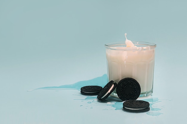 Splashes of milk in a glass, next to it lies a chocolate chip cookie on a light blue background.