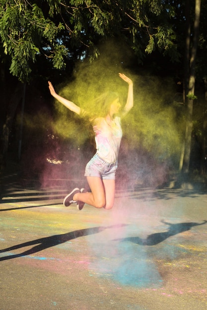 Splendid brunette woman jumping with exploding pink, blue and yellow Holi powder in the park