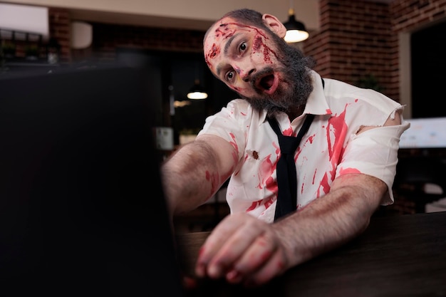 Spooky evil zombie using laptop at desk, looking creepy and horrific working on computer. Terrifying eerie dramatic brain eating monsters sitting in business office, horrible gory walker.