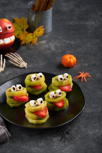Spooky green kiwi monsters for Halloween Healthy Fruit Halloween Treats Halloween party kiwi strawberry apple and marshmallow monster on dark grey stone or concrete table background