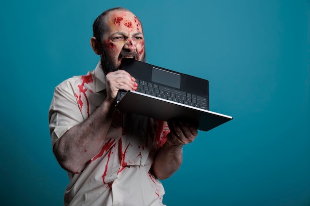 spooky halloween zombie biting on laptop computer, having bloody scars and wounds on creepy dirty face. frightening undead monster being aggressive with pc, looking dangerous cruel and deadly.