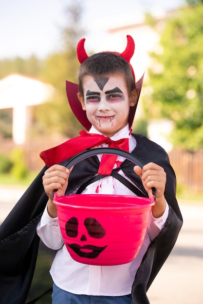 Photo spooky little boy in halloween costume of devil with horns on head holding basket with treats and looking at you against country houses