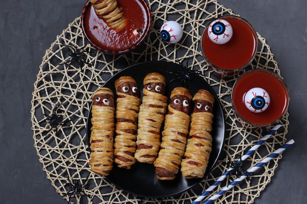 Spooky sausage mummies, tomato juice and sauce for Halloween party on black. Top view.