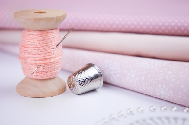A spool of thread a needle a thimble and pink fabrics on a white table