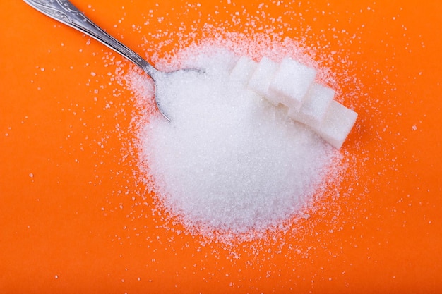 Spoon and cubes of white sugar and granulated sugar on an orange background