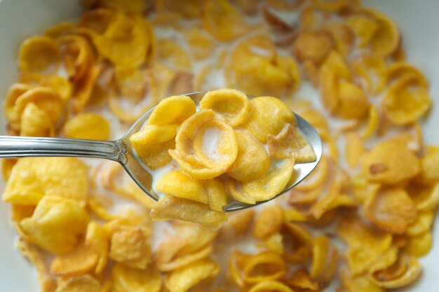 Spoon of delicious crispy cornflakes with milk above plate closeup