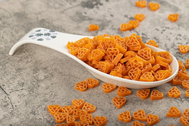 Spoon of raw heart-shaped pasta scattered on stone background. 