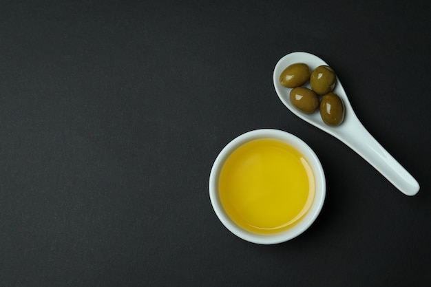 Spoon with olives and bowl of oil on black surface