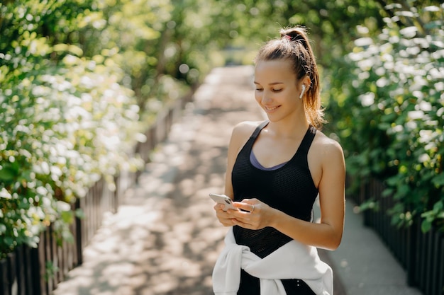 Sport als levensstijl Horizontaal schot van gezonde slanke sportieve vrouw gefocust op smartphone controleert informatie en kiest nummer uit afspeellijst gekleed in sportkleding poseert buiten ademt frisse lucht