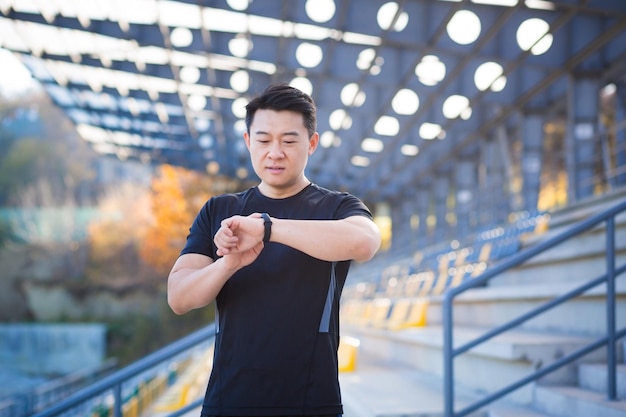 Спортивный азиатский мужчина смотрит на умные часы на современном треке Бегун с браслетом SmartWatch Портрет фитнес-мужчины проверяет результат Мужчина в спортивной одежде бегает трусцой по улице на городском фоне городской стадион