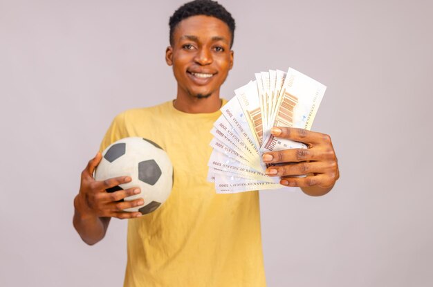 Sport bets portrait of excited black guy with football ball and
money standing over beige background