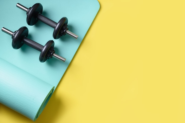 Sport black dumbbells on turquoise yoga mat on yellow. Copy space. View from above.