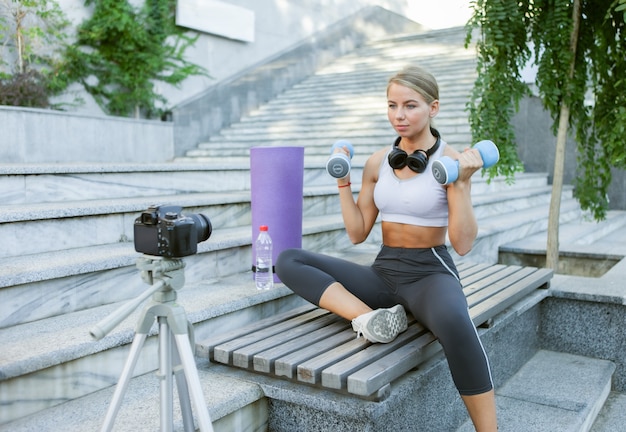 Sport blog. Attractive young sports woman working out with dumbbells outdoor, demonstrating exercises for her blog, records on camera on tripod. Lesson for her fitness vlog