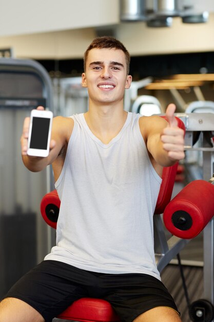 спорт, бодибилдинг, образ жизни, технологии и концепция людей - улыбающийся молодой человек показывает смартфон и показывает большой палец вверх в спортзале