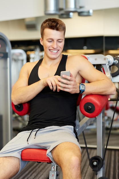 спорт, бодибилдинг, образ жизни, технологии и концепция людей - улыбающийся молодой человек со смартфоном в спортзале