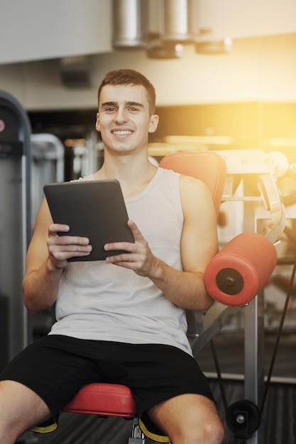 sport, bodybuilding, lifestyle, technology and people concept - smiling young man with tablet pc computer in gym