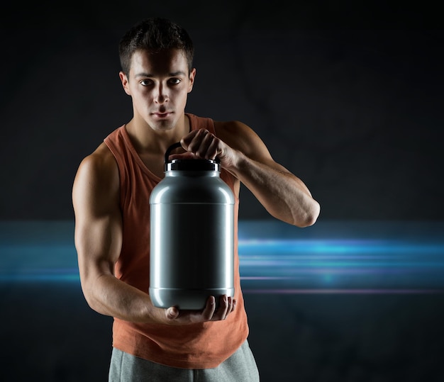 sport, bodybuilding, strength and people concept - young man standing holding jar with protein over dark background