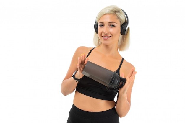 Sport caucasian blonde girl stands with earphones do sports and drinks water from sport bottle that she show