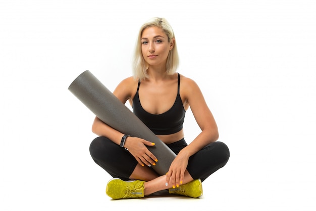 Sport caucasian girl sits on floor, cross legs and keeps a gymnastic mat isolated