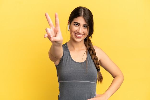 Sport donna caucasica isolata su sfondo giallo sorridente e mostrando segno di vittoria