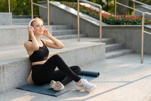 Sport en fitness Jonge volwassen blanke vrouw Zitten Trainingsmat Betonnen vloer Rusten na ochtend Training Buiten Zomer Park Atletisch Vrouw Pauze luisteren Muziek Hoofdtelefoons